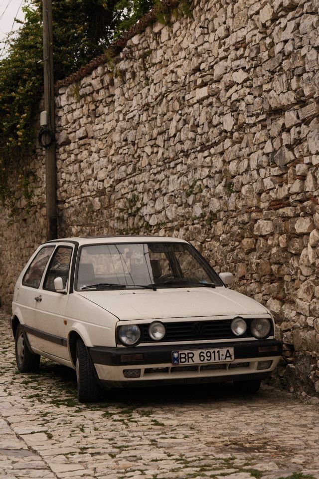 Un coche clásico blanco estacionado en una calle empedrada junto a un alto muro de piedra rústico.