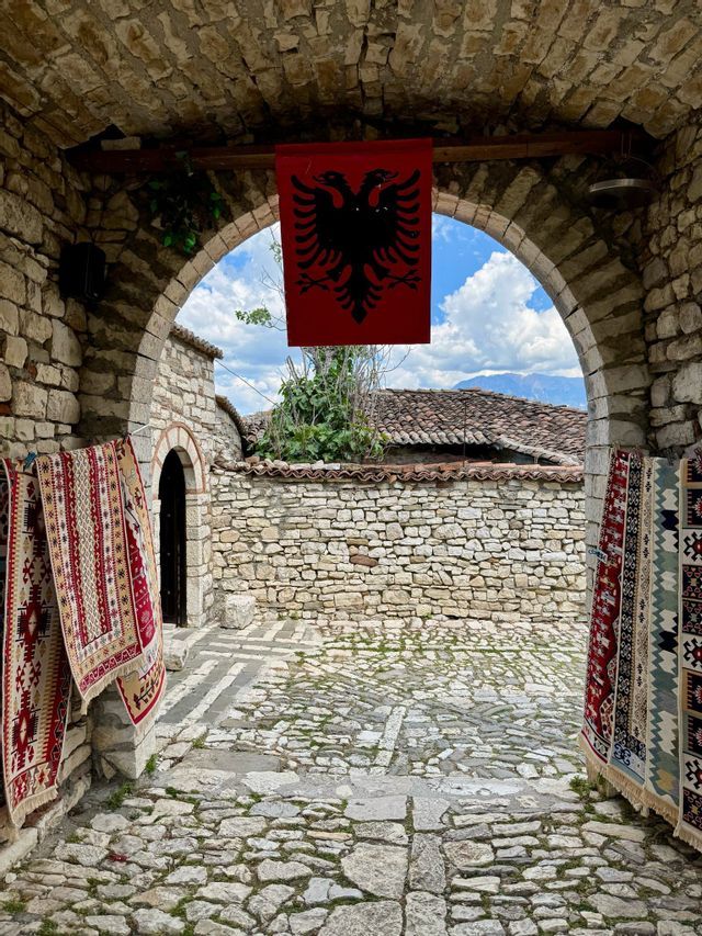 An Albanian flag hangs from a stone archway over a cobblestone courtyard, with traditional rugs for sale on the walls.