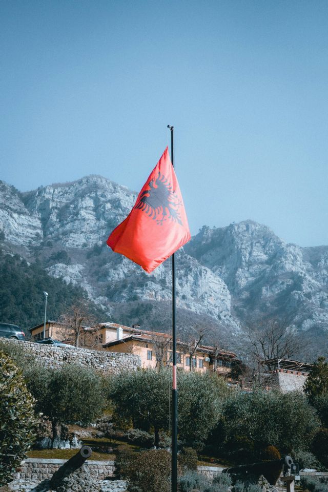 La bandera albanesa ondea en un mástil frente a una gran cordillera rocosa bajo un cielo despejado.