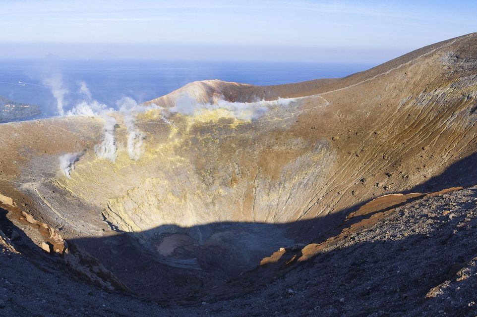 Ampia vista di un cratere vulcanico con depositi di zolfo giallo e vapore che sale dalle fumarole. Il mare è visibile in lontananza.