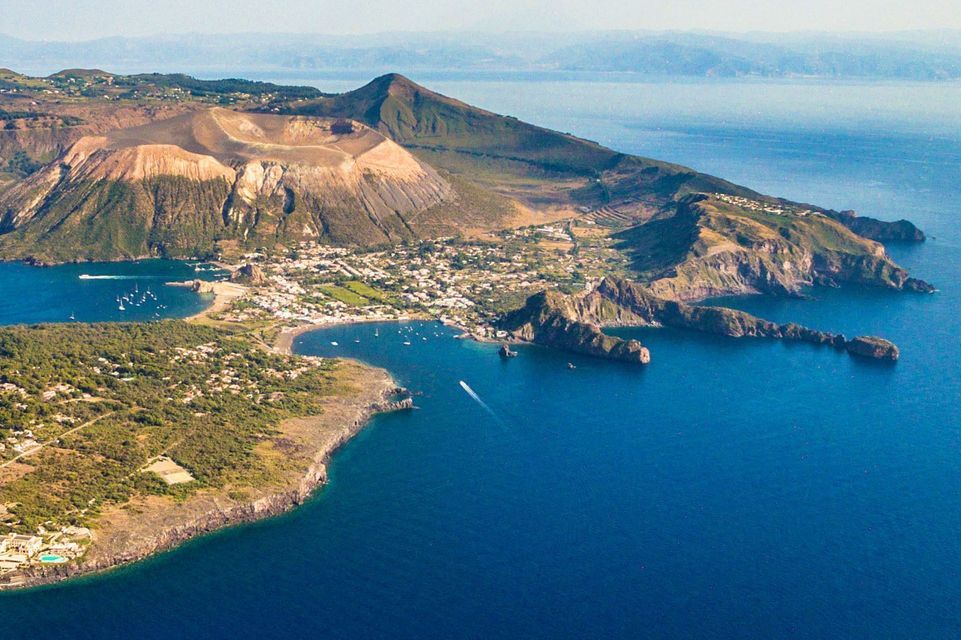Veduta aerea di un'isola vulcanica caratterizzata da un ampio cratere, con un paese e imbarcazioni in una baia lungo la costa.
