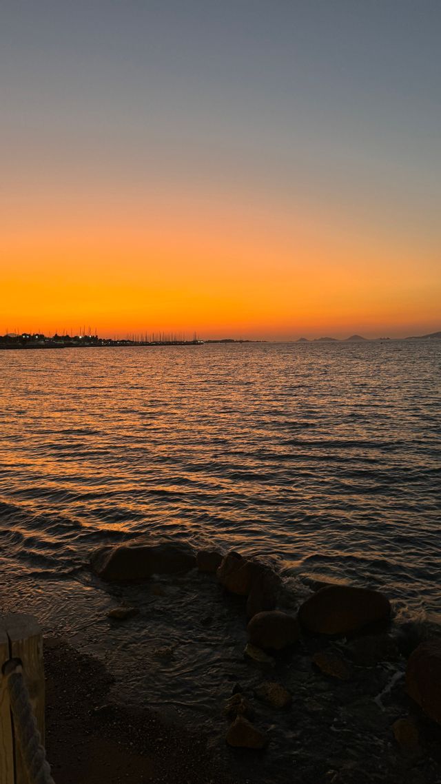 Il mare riflette un tramonto arancione vibrante, con la sagoma di un porto lontano all'orizzonte.