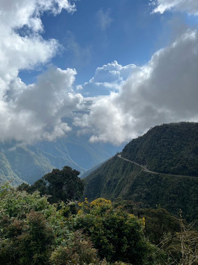Una strada stretta si snoda lungo una montagna verde e lussureggiante, affacciandosi su una valle sotto un cielo azzurro parzialmente nuvoloso.