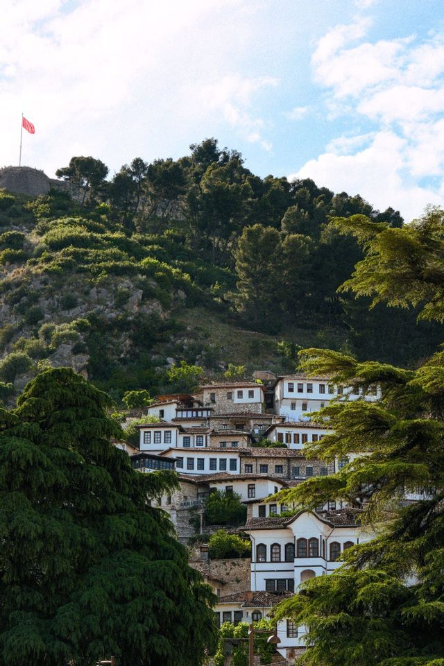 Casas blancas tradicionales con techos de piedra se agrupan en una exuberante ladera verde debajo de una fortaleza con una bandera roja.