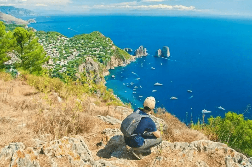 Una persona con una mochila se agacha en la cima de un acantilado rocoso, mirando hacia un mar azul brillante salpicado de barcos y un pueblo costero.