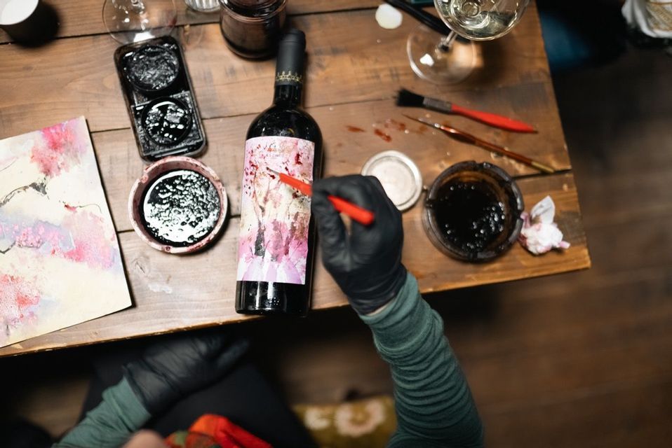 Vista dall'alto di una persona con guanti neri che dipinge l'etichetta di una bottiglia di vino su un tavolo di legno, circondata da materiali d'arte.