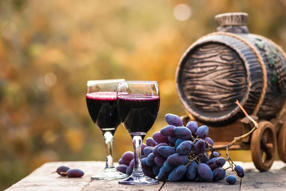 Due bicchieri di vino rosso, un grappolo d'uva viola e una botte decorativa su un tavolo di legno rustico all'aperto.