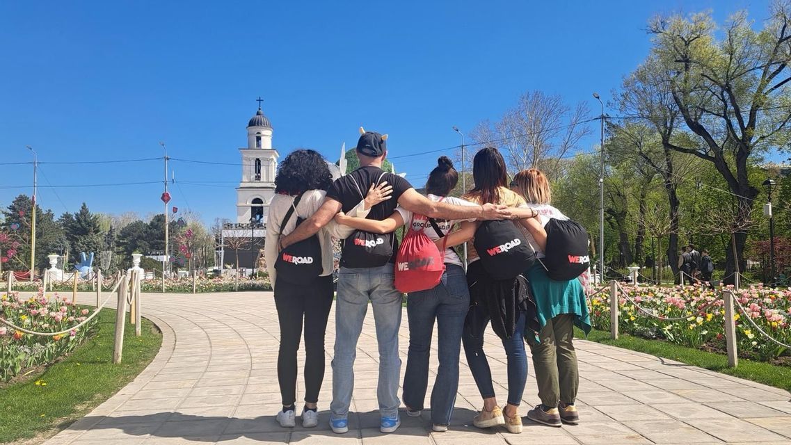 Un gruppo WeRoad di sei persone si abbraccia in un parco, di fronte a un lontano campanile bianco.
