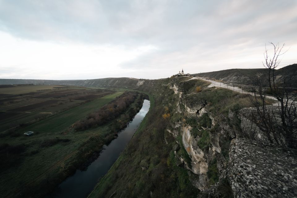 Un fiume serpeggia in una valle, accanto a un'alta scogliera rocciosa su cui si erge una piccola chiesa, sotto un cielo coperto.