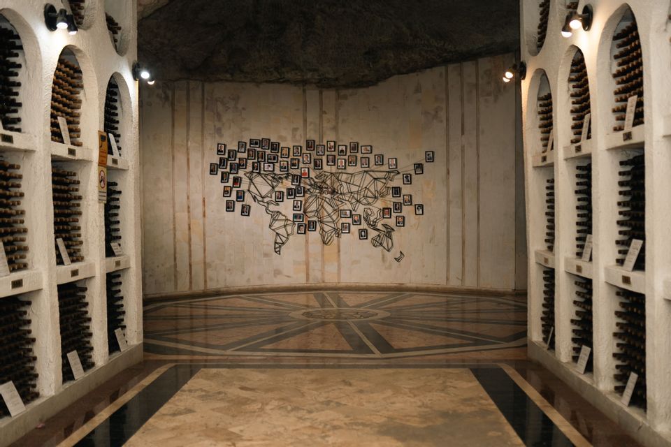 Una cantina con scaffali ad arco pieni di bottiglie e una grande mappa del mondo fatta di foto sulla parete di fondo.