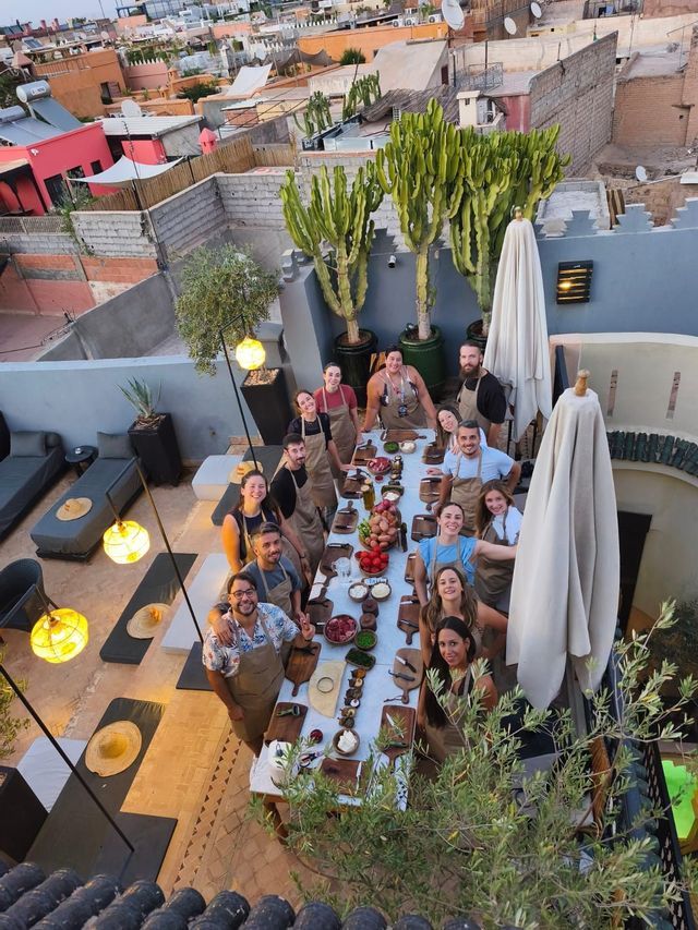 Un viaje en grupo de WeRoad, con delantales, posa alrededor de una larga mesa preparada para una clase de cocina en una terraza en la azotea con vistas a una ciudad.