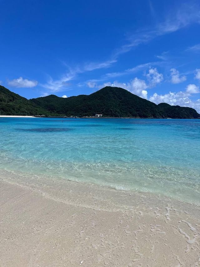 Acqua cristallina color turchese su una spiaggia di sabbia bianca con montagne verdi ricoperte di alberi in lontananza sotto un cielo blu.