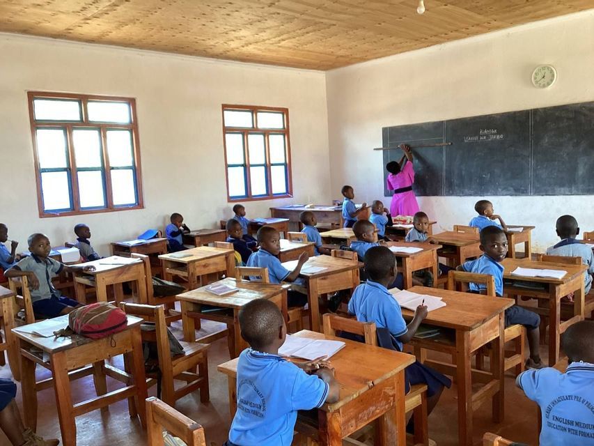 Una maestra in un vestito rosa scrive su una lavagna mentre giovani studenti in uniformi blu osservano dai loro banchi di legno in un'aula.