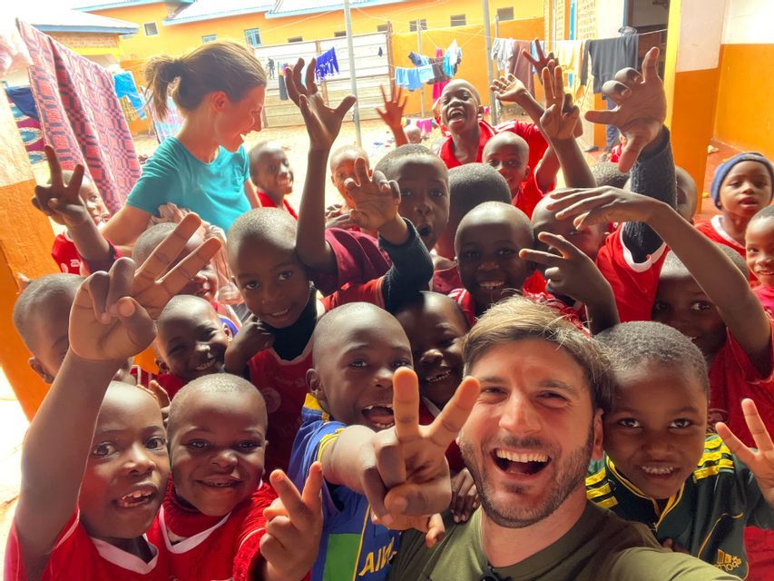 Due viaggiatori WeRoad si scattano un selfie di gruppo con tanti bambini sorridenti che fanno il segno della pace in un cortile esterno.