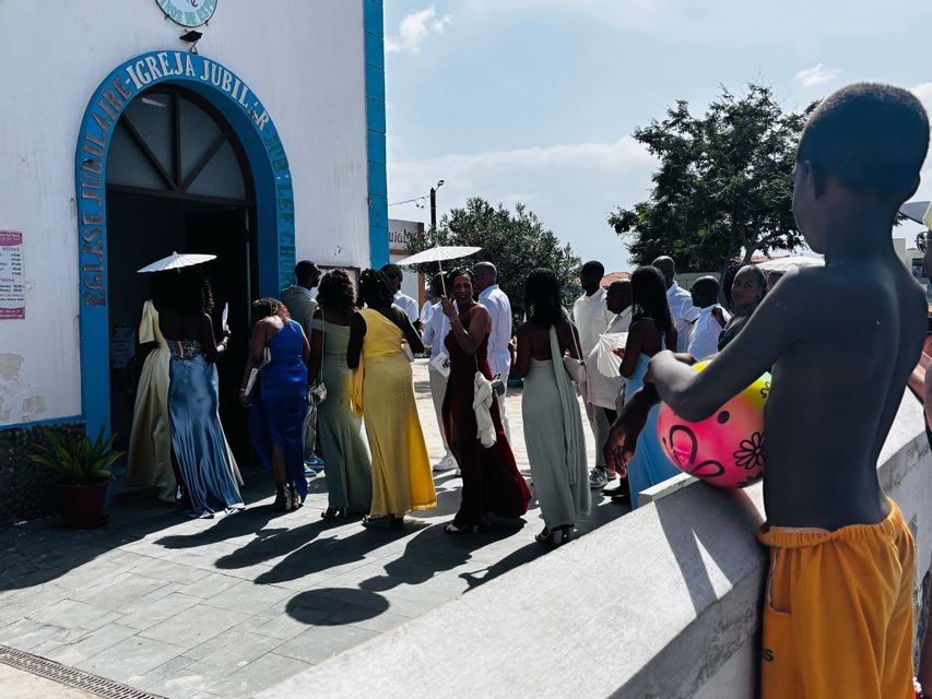 Un gruppo di persone in abiti formali e colorati si riunisce all'esterno di una chiesa in una giornata di sole, mentre un ragazzo in primo piano osserva.