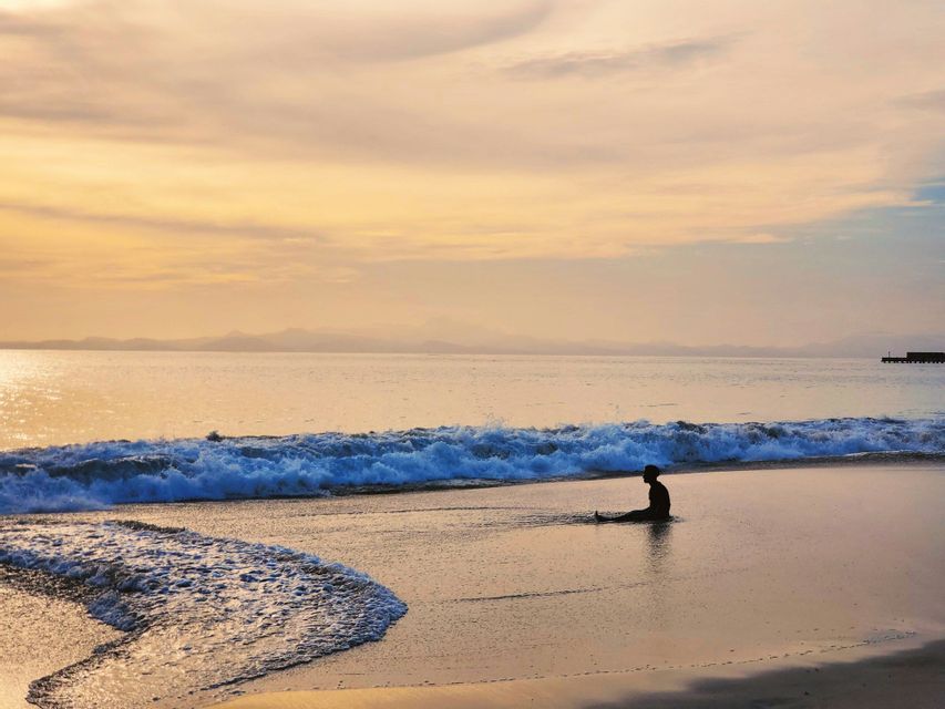 Una silhouette di una persona seduta tra le onde basse su una spiaggia sabbiosa durante un tramonto dorato.