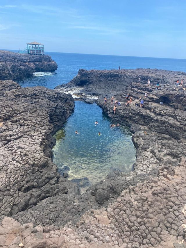 Una vista dall'alto di un gruppo WeRoad che nuota e si rilassa in una piscina naturale di roccia sull'oceano in una giornata di sole.