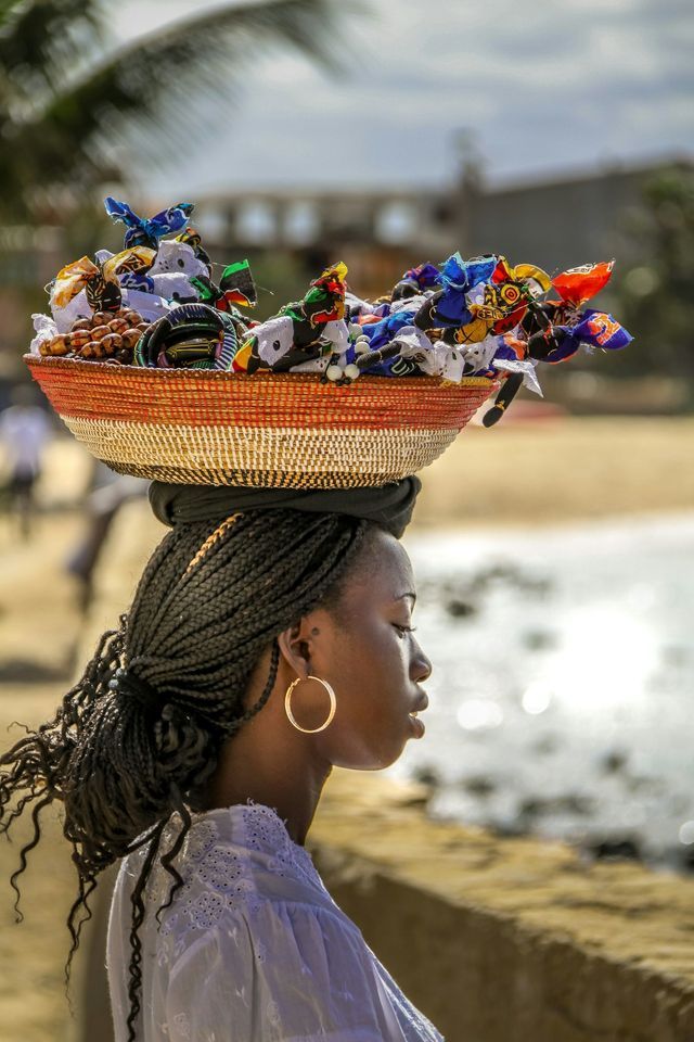 Una donna di profilo, con capelli intrecciati, bilancia sulla testa un cesto intrecciato ricolmo di artigianato colorato, vicino a una spiaggia.