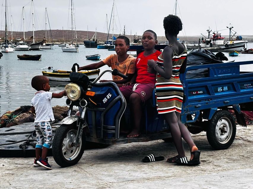 Quattro bambini si radunano attorno a un triciclo motorizzato blu su un molo, con un porto pieno di barche sullo sfondo.