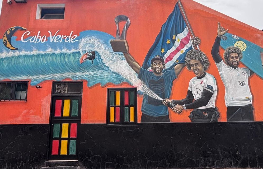 Un murale colorato su un edificio arancione raffigura tre atleti che celebrano una vittoria negli sport acquatici con un trofeo e la bandiera di Capo Verde.