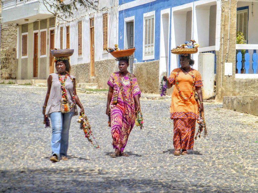 Tres mujeres con ropa colorida caminan por una calle empedrada, equilibrando cestas de artesanías sobre sus cabezas.
