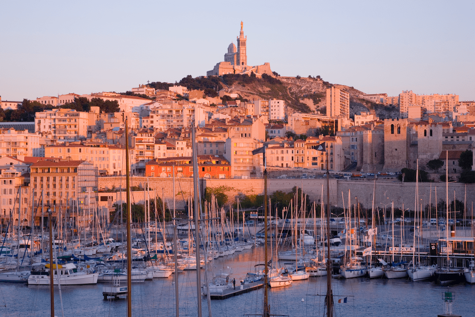 A harbor full of sailboats at the base of a hillside city, with a basilica on the hilltop, seen during a warm sunset.