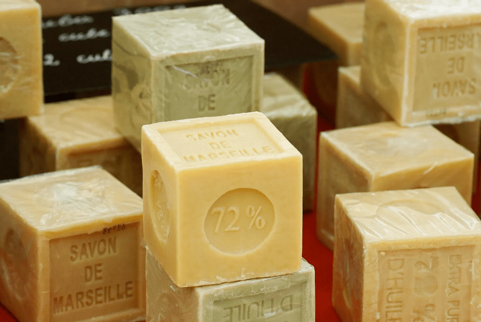A close-up of stacked, cube-shaped bars of Savon de Marseille soap. One unwrapped bar in the foreground is stamped with '72%'.