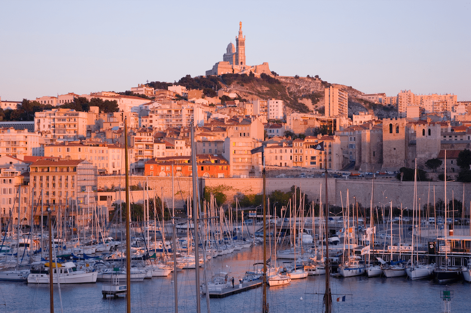 Sailboats docked in a harbor at sunset, with a city and a basilica on a hill in the background.