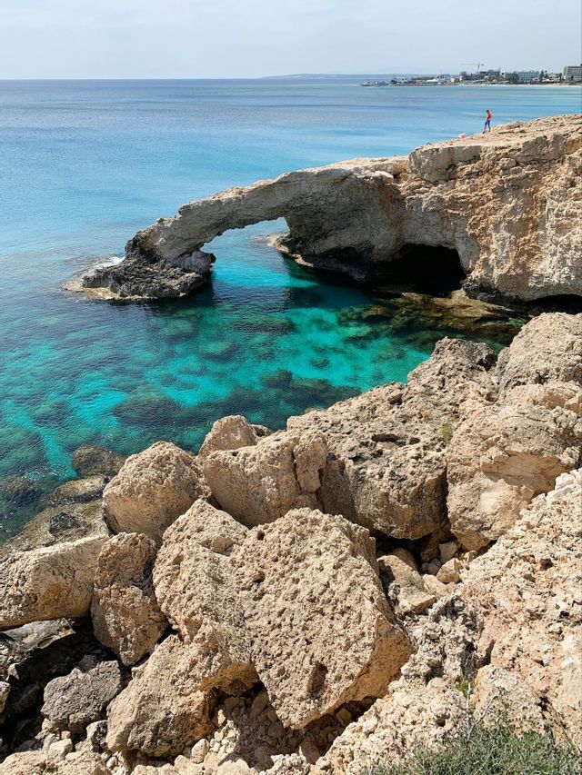 Un arco di roccia naturale su una costa frastagliata si protende su acque limpide e turchesi sotto un cielo soleggiato.
