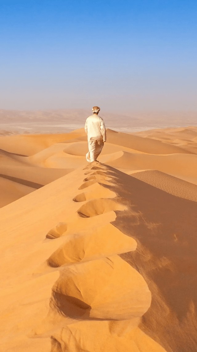 A person in traditional attire walks along the crest of a large sand dune in a vast desert, leaving footprints behind them.