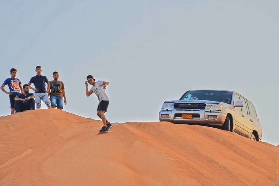 A man from a WeRoad group trip sandboards down a large desert dune as his friends watch from the top, next to a silver SUV.
