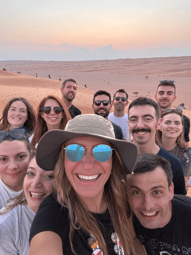 A woman in a hat takes a selfie with her WeRoad group trip on a vast sand dune at sunset.