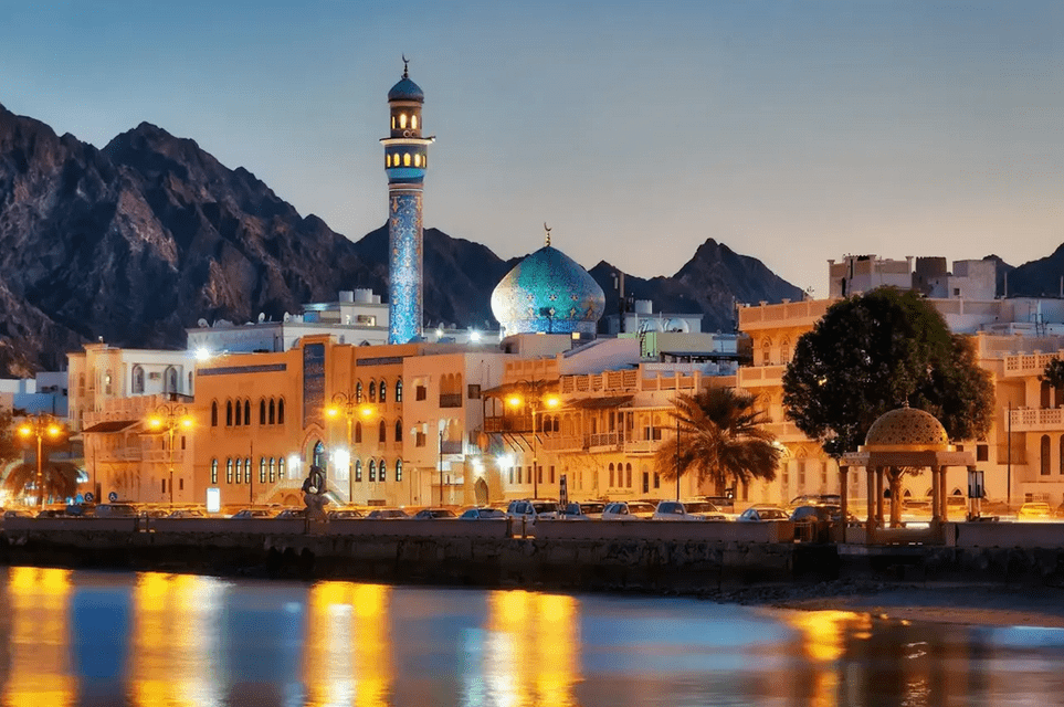 An illuminated city waterfront at dusk, featuring a mosque with a blue dome and minaret set against a backdrop of dark mountains.