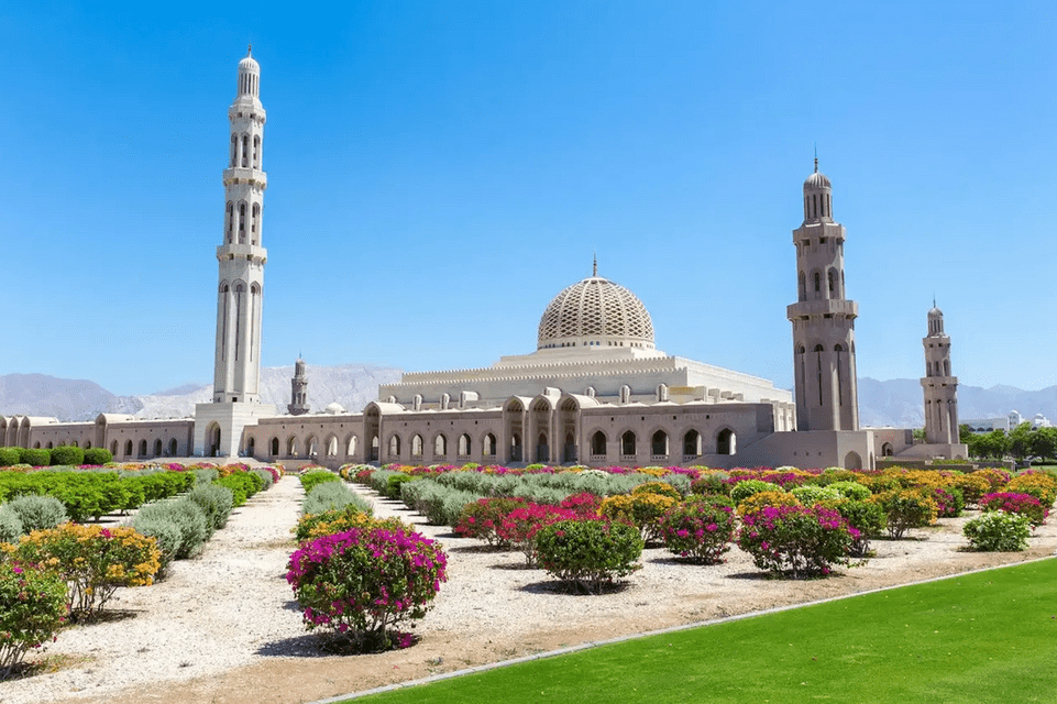 A large, light-colored mosque with a patterned dome and tall minarets, set behind a manicured garden with rows of colorful flowering bushes.