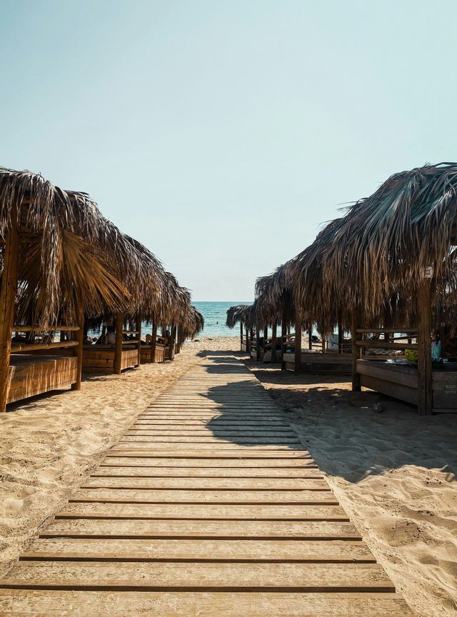 Una passerella di legno su una spiaggia sabbiosa conduce tra file di cabine con tetti di paglia verso l'oceano.