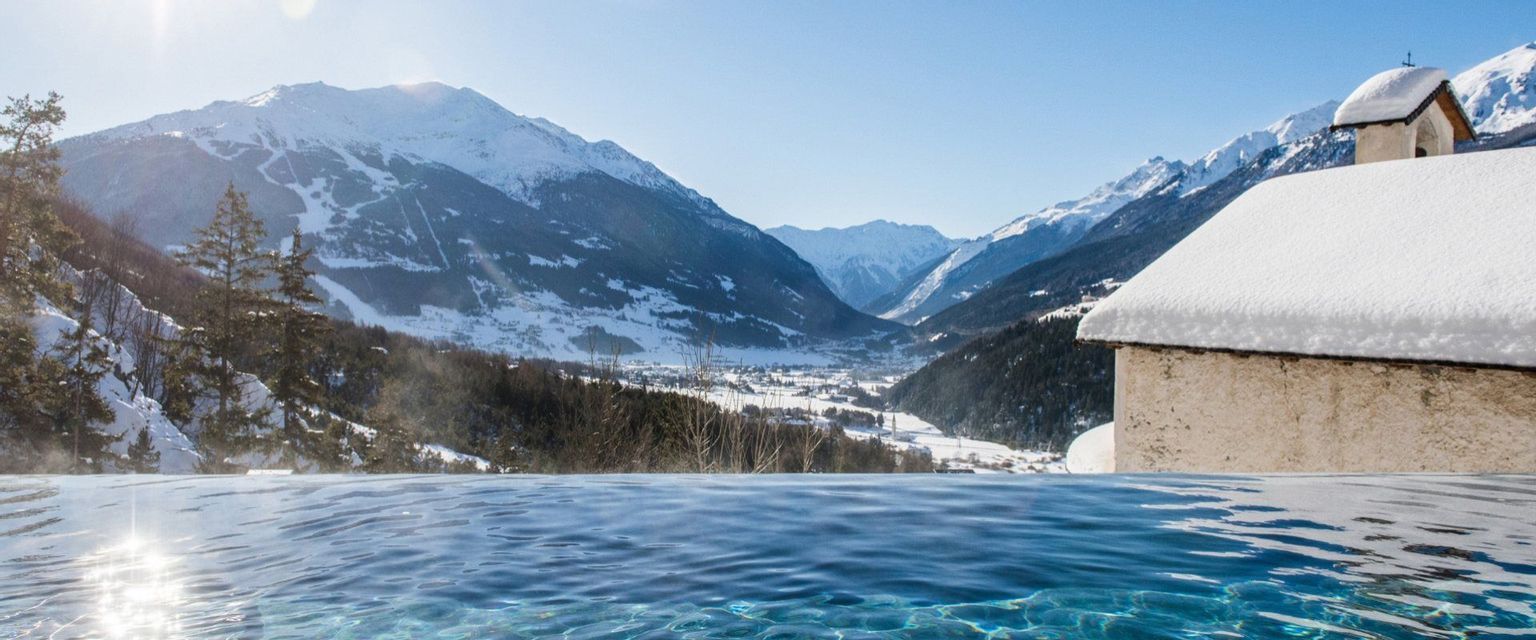 Una piscina a sfioro si affaccia su una valle soleggiata e montagne innevate, con un edificio innevato sulla destra.
