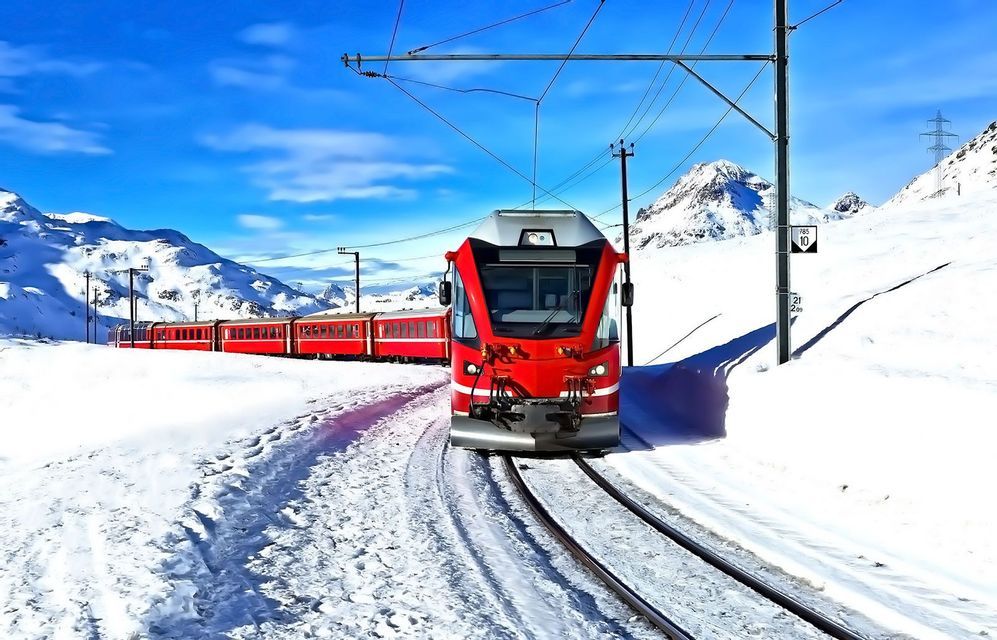 Un treno rosso viaggia sui binari attraverso un paesaggio montano innevato sotto un cielo azzurro chiaro.