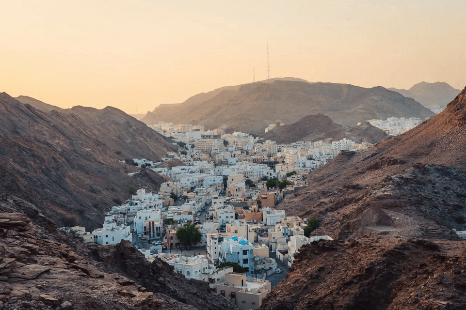 A city of white buildings nestled in a valley between arid, rocky mountains during a golden sunset.