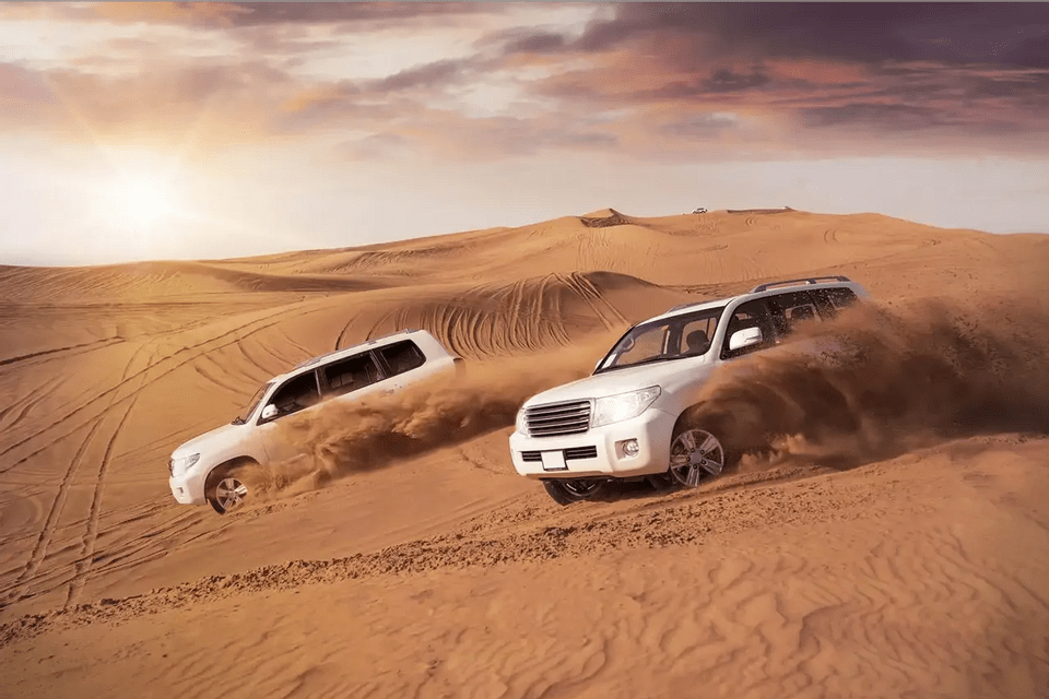 Two white SUVs kick up sand while driving down a steep dune in the desert at sunset.
