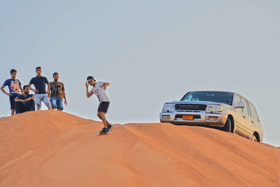 A person from a WeRoad group trip sandboards down an orange dune as others watch from the top next to a silver SUV.