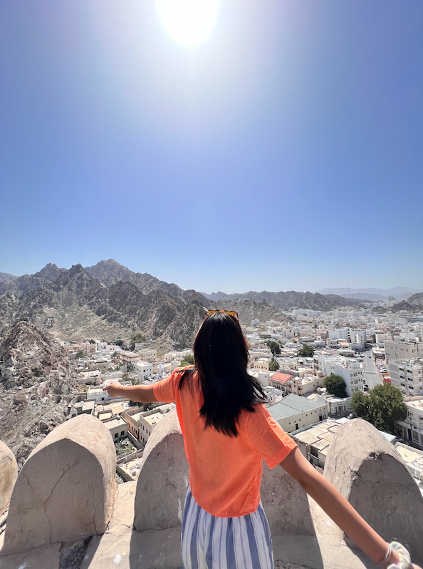 A woman in an orange shirt stands with arms outstretched, looking over a city nestled between mountains under a bright, sunny sky.