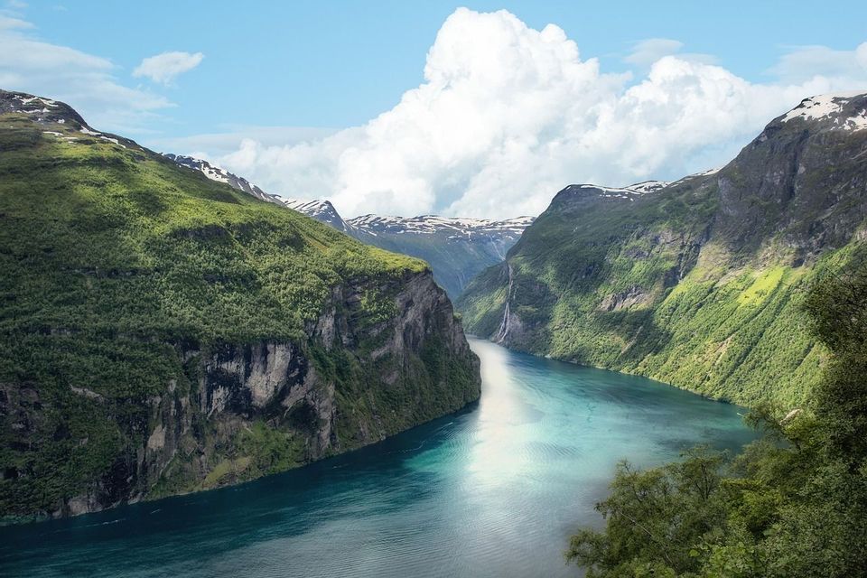A turquoise fjord winds between steep, forest-covered mountains with snow-capped peaks under a blue sky with white clouds.