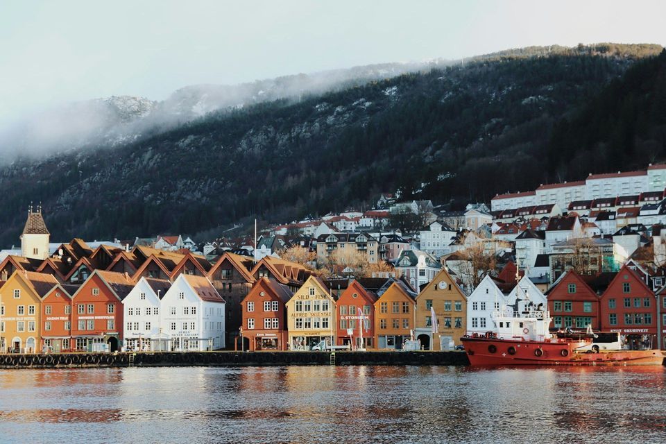 A row of colorful wooden houses lines a waterfront, with a red boat moored alongside and a forested hill rising in the background.