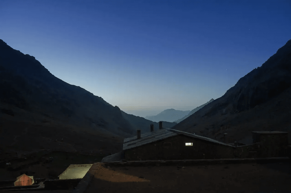 A rustic building with one lit window sits in a mountain valley at dusk under a deep blue sky.