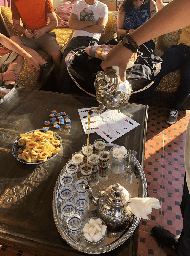 Une personne verse du thé traditionnel d'une théière argentée dans des verres pour un groupe WeRoad réuni autour d'une table avec des biscuits.