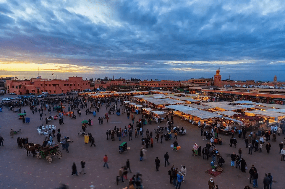 Ein belebter Stadtplatz in der Dämmerung, von oben gesehen, voller Menschen und beleuchteter Marktstände.