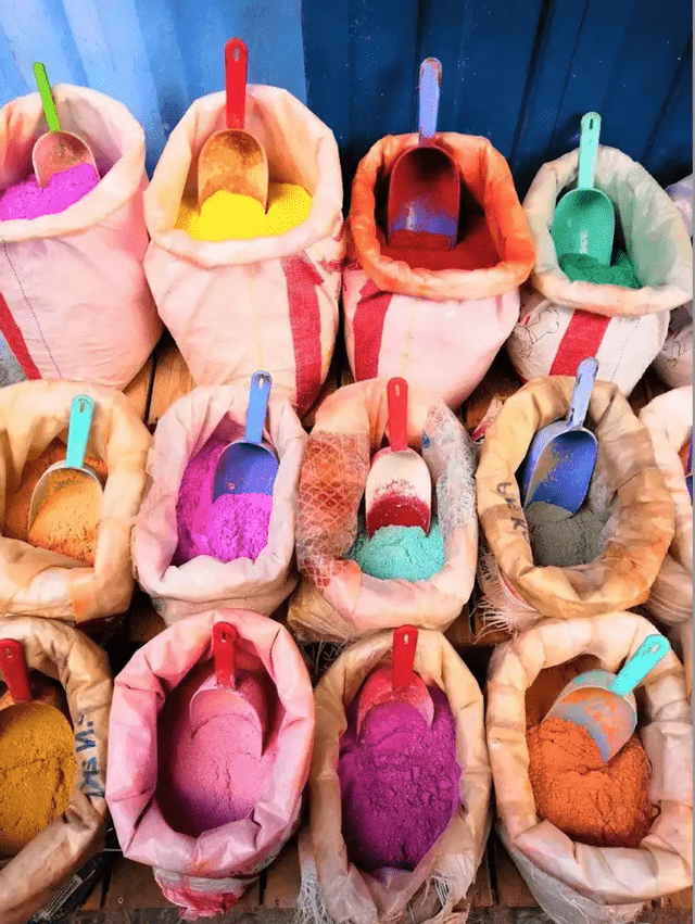 An overhead view of numerous open sacks filled with vibrant powdered pigments, each containing a colorful plastic scoop.