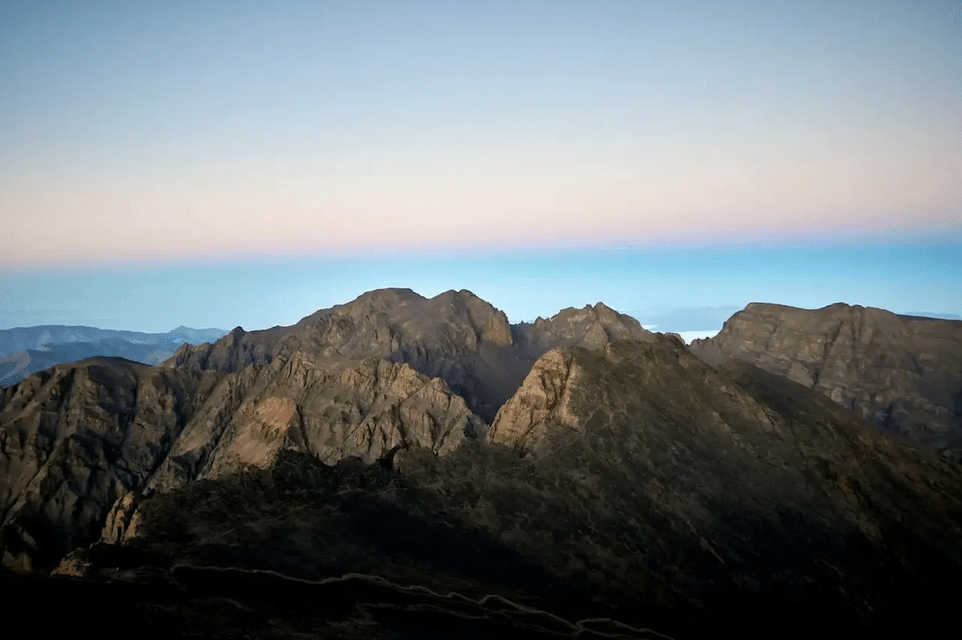 Rauhe, dunkle Berggipfel heben sich gegen einen klaren Himmel mit einem rosa-blauen Farbverlauf am Horizont im Morgengrauen ab.