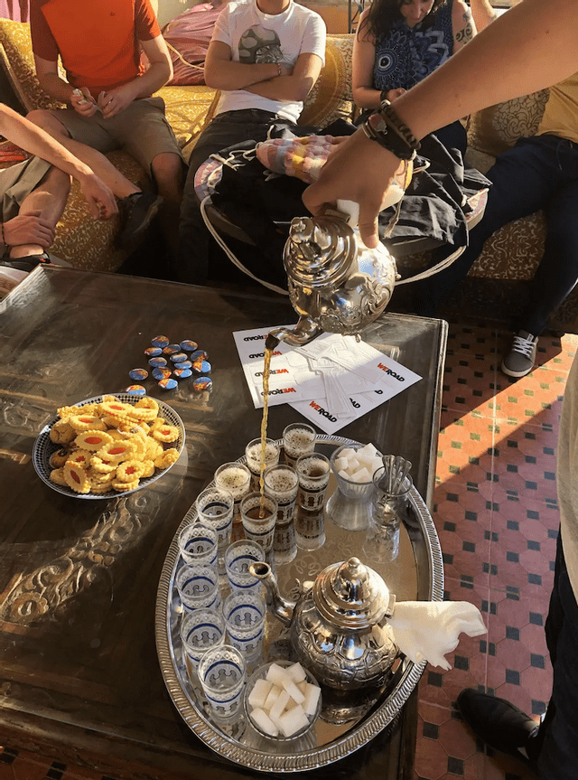 A person pours tea from an ornate silver teapot into small glasses for a WeRoad group trip, with cookies on the table.