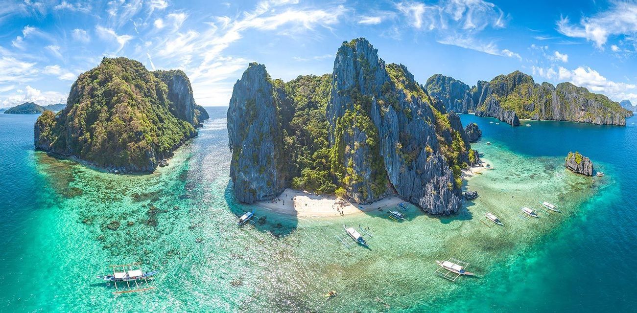 Vista aerea di isole rocciose e ricoperte di vegetazione, con barche a bilanciere su acque limpide e turchesi, vicino a una piccola spiaggia sabbiosa.